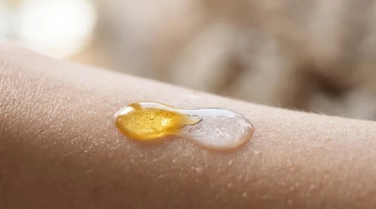 Gros plan artistique sur des gouttes de serum nettoyant doré fusionnant avec de l'eau pure sur une peau lumineuse, evoquant la purete alpine